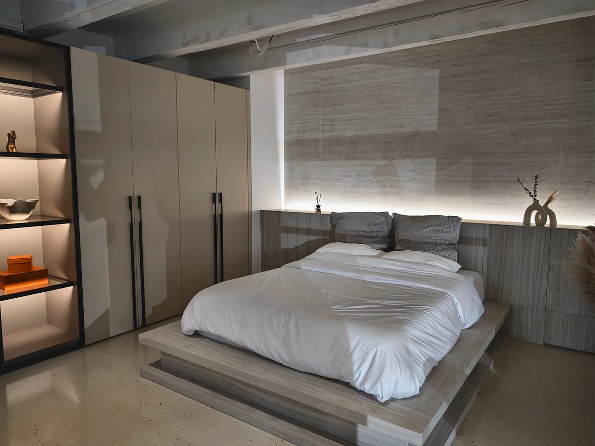 A staged bed with white linen against a lit stone wall, with fitted wardrobes and shelving beside it.
