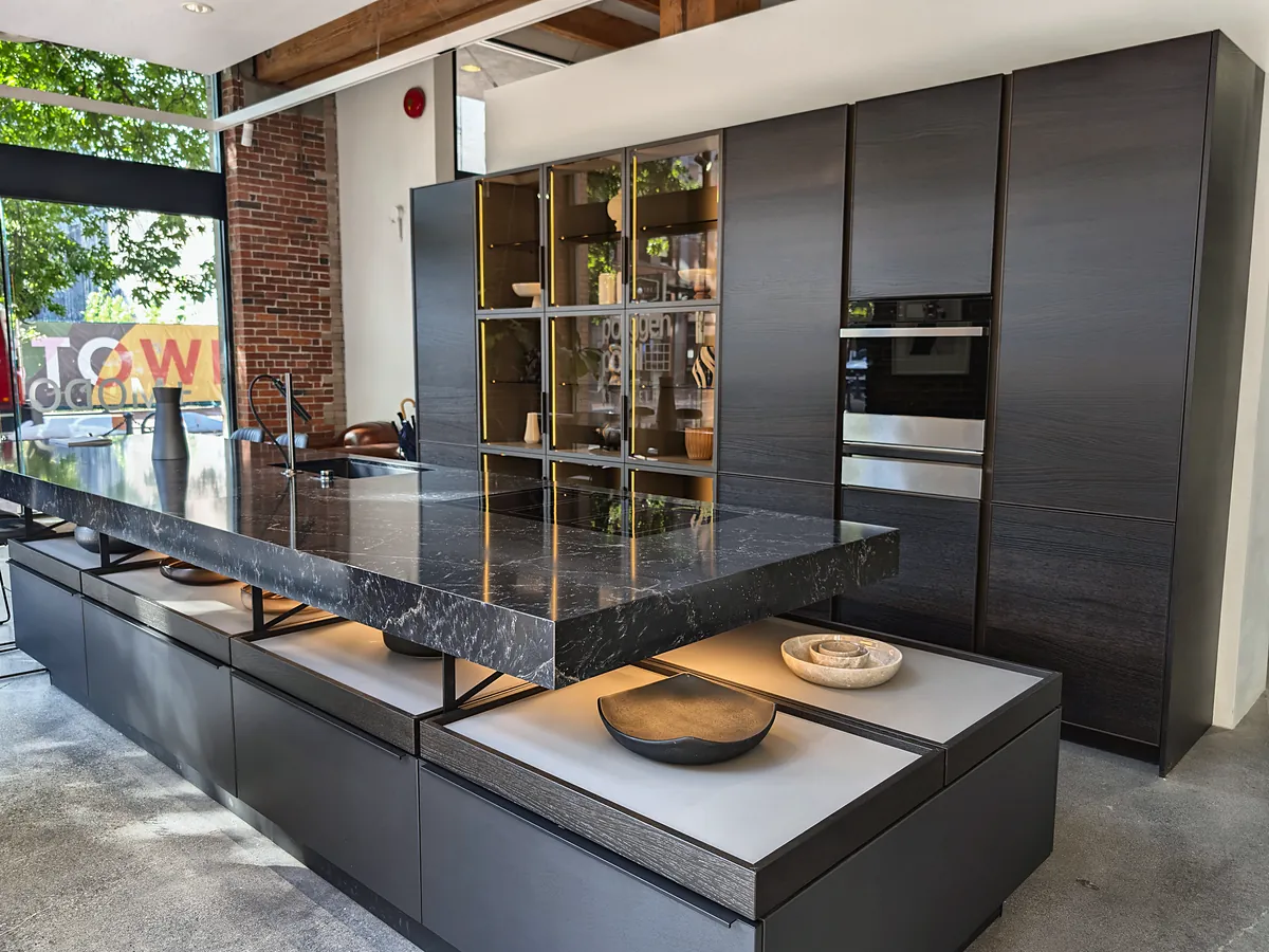 A black stone worktop floating above lit shelving, with dark tall cabinets and glass doors behind.