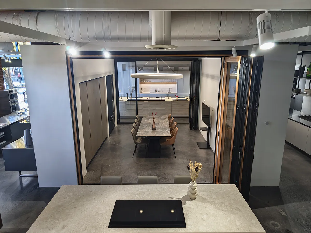 View through open folding glass doors into a dining room with a long table and a kitchen behind it.