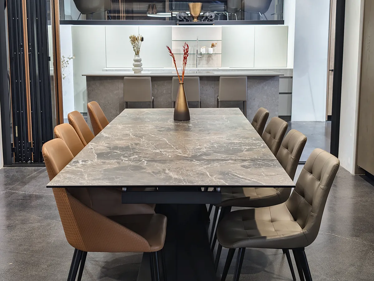 A long stone-topped dining table with tan and taupe leather chairs and a bronze vase.