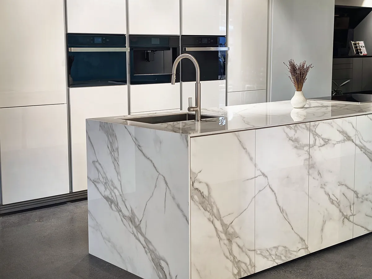 A white marble waterfall island in front of a white wall of ovens and a coffee machine.