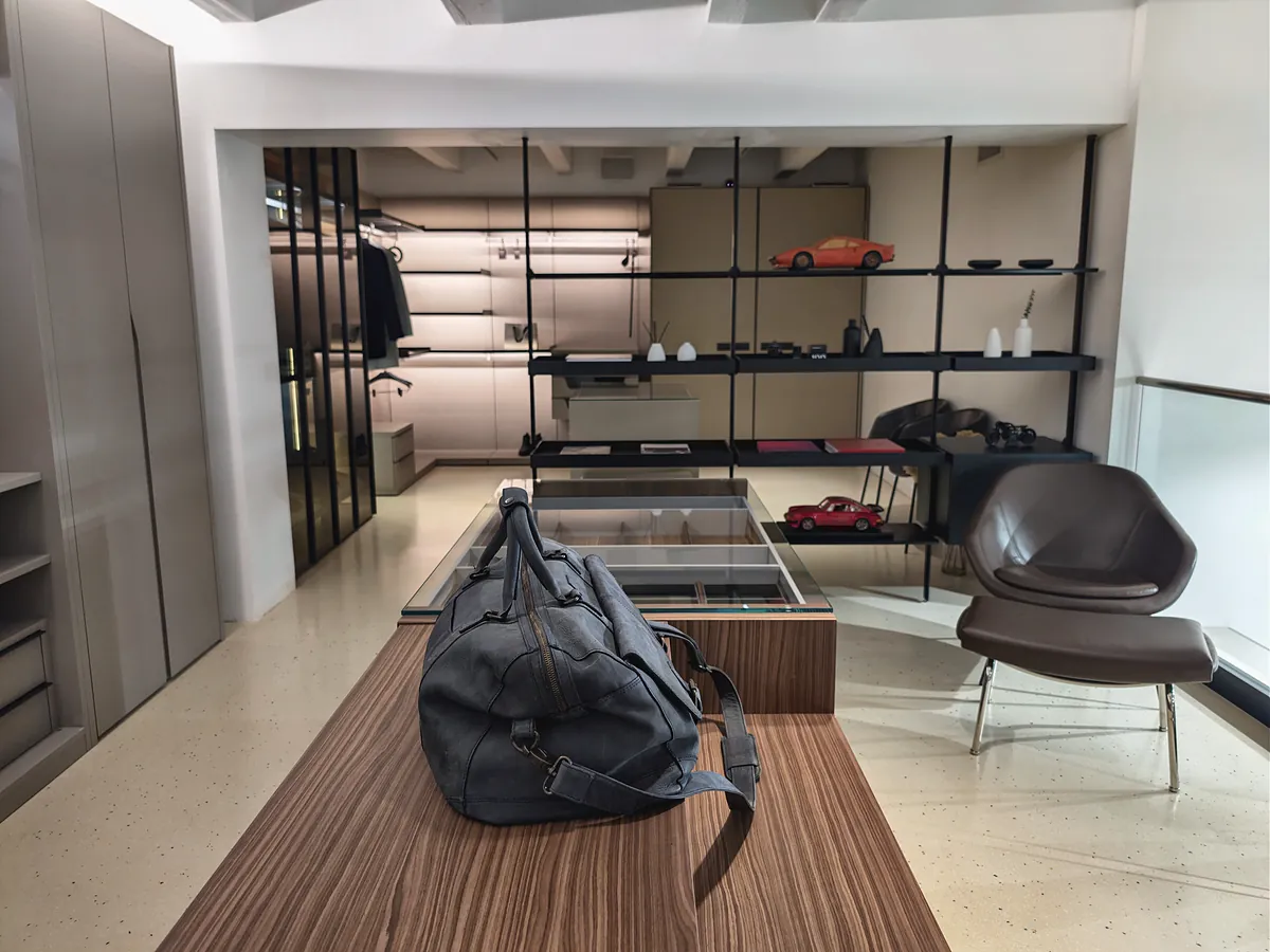 A walk-in wardrobe behind glass with a walnut dressing island, a leather chair and a mezzanine guardrail.